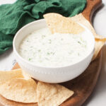 Crockpot spinach dip in a white bowl surrounded by tortilla chips. There is a green linen in the background.