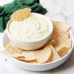 Creamy Green Avocado Dip surrounded by tortilla chips with a green tea towel in the background.