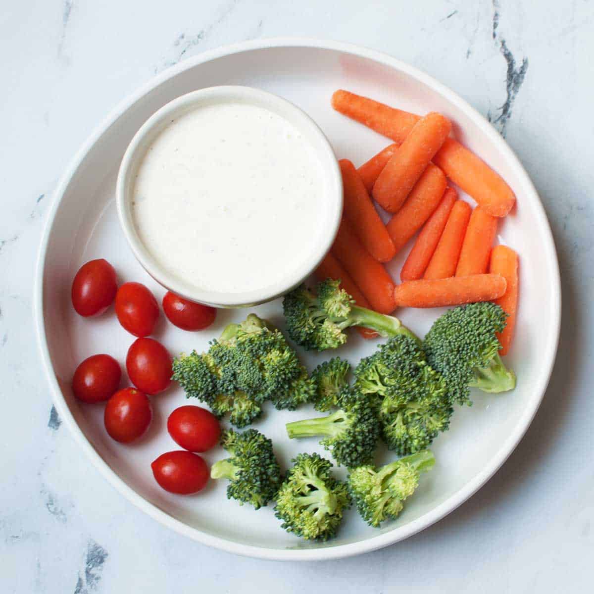 bowl of ranch surrounded by carrots, broccoli, and tomatoes.