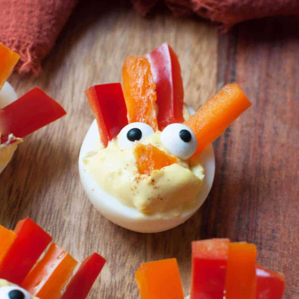 a close up view of a deviled egg that has candy eyes and bell pepper strips arranged to look like a turkey