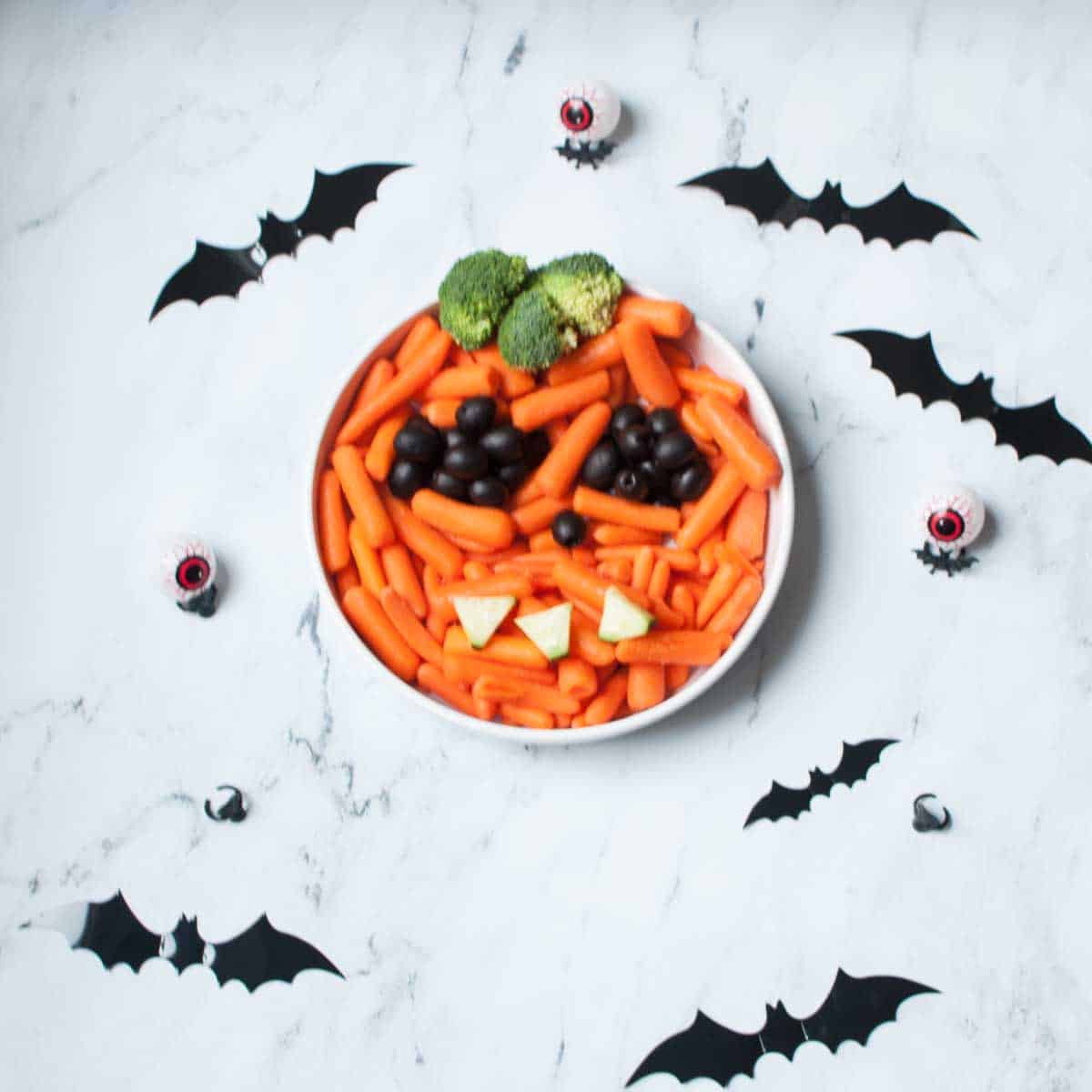 A halloween veggie tray. There are carrots in a dish with olives, broccoli, and cucumbers. They are arranged in a way that makes it look like a jack-o-latern. There are bat and eyeball Halloween decorations surrounding it.