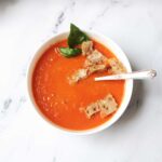 copycat panera tomato soup with a spoon, basil leaf, and croutons