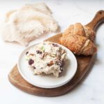 Chicken salad with cranberries on a white plate next to croissants on a wooden tray.