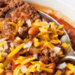 Bison chilil with cheese and onions on top. There is a large spoon scooping it.