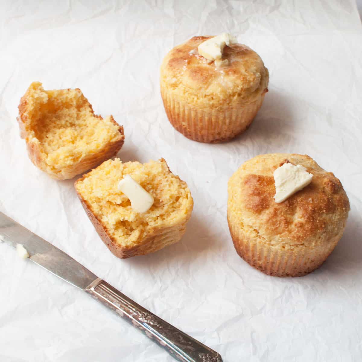 Jumbo corn muffins with butter on top on a white background. There is a butterknife with butter beside them.