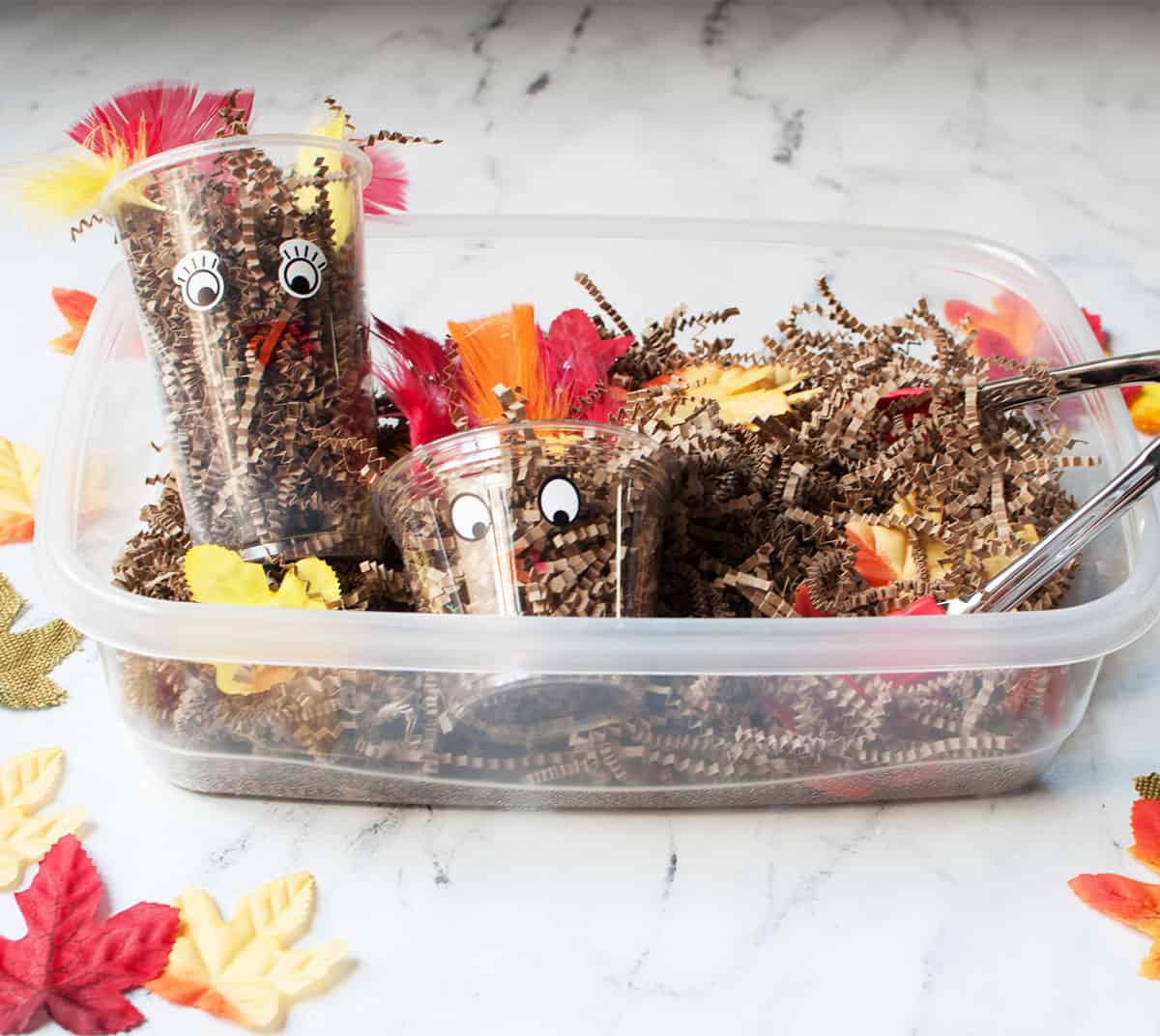 A turkey sensory bin made from plastic cups with turkey faces drawn on them and brown crinkle paper.
