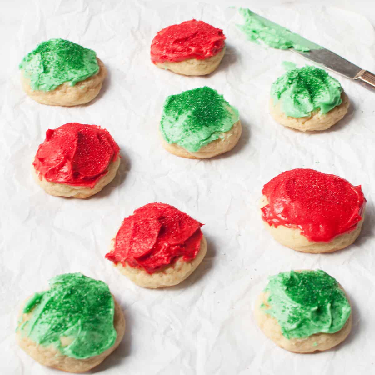 Amish Christmas cookies with green or red frosting and sprinkles on a white backbground. There is a knife with frosting on it in the background.
