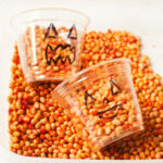 A pumpkin themed sensory bin meant for toddlers and preschoolers. There are small beans that have been dyed orange and cups with pumpkin faces on them.