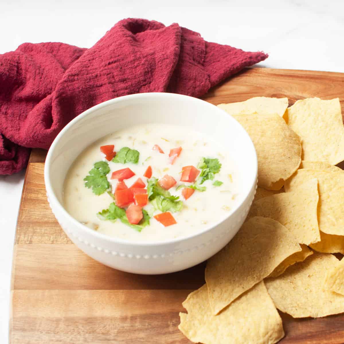White queso (cheese dip) in a white bowl with cilantro and tomato on top. There are tortilla chips and a red linen around it.