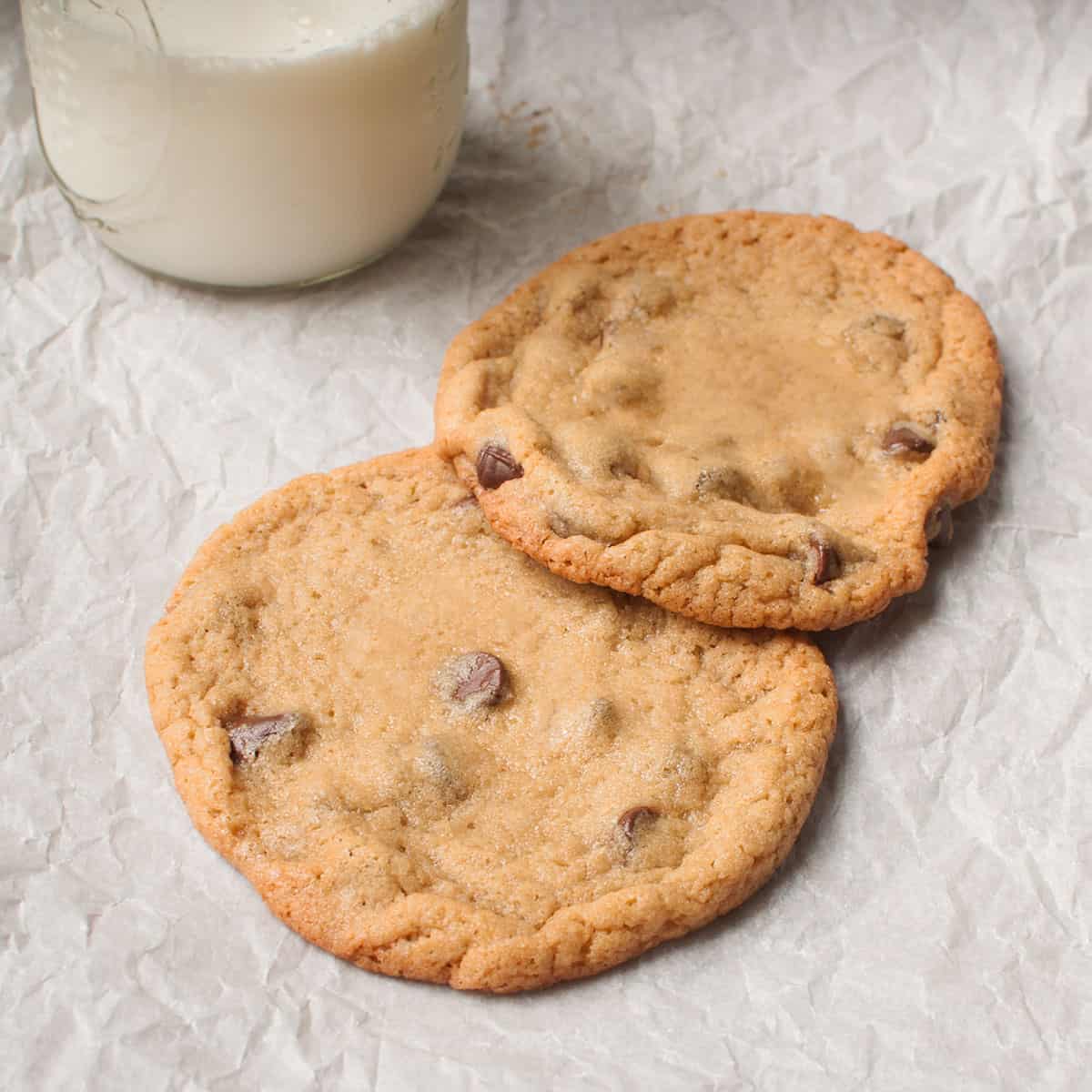 Two very large chocolate chip cookies on a white background with a glass of milk.