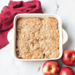 Sourdough apple crisp in an 8 inch casserole dish. There are apples and a red linen beside it.