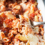 A spoon scooping baked ziti with ground turkey from a white casserole dish.