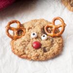 An oatmeal cookie made to look like reindeer with candy eyes and pretzels on a white background.