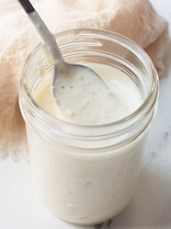Copycat Carrabba's creamy parmesan salad dressing in a mason jar with a spoon in it. There is a cream linen behind it.