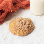 A photograph of a pumpkin muffin cookie on a white background. There is an orange linen and a cup of milk in the background.