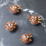 Four Halloween muffins which are chocolate banana muffins with candy eyes and pretzel sticks made to look like spiders.