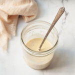 A mason jar filled with Copycat Texas Roadhouse Italian Dressing. There is a spoon sticking out of the jar and a cream linen.