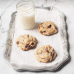 Very large and thick chocolate chip cookies on a silver tray with white parchment paper on it. There is a glass of milk behind the cookies.