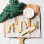 Copycat Carrabba's zucchini on a wooden board with garlic aioli sauce in a white dish. There is a green linen above the zucchini.