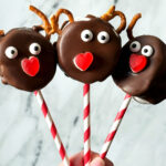 The very top of a hand holding three chocolate covered marshmallows made to look like reindeer. The treats have candy eyes, pretzels, and a candy nose.