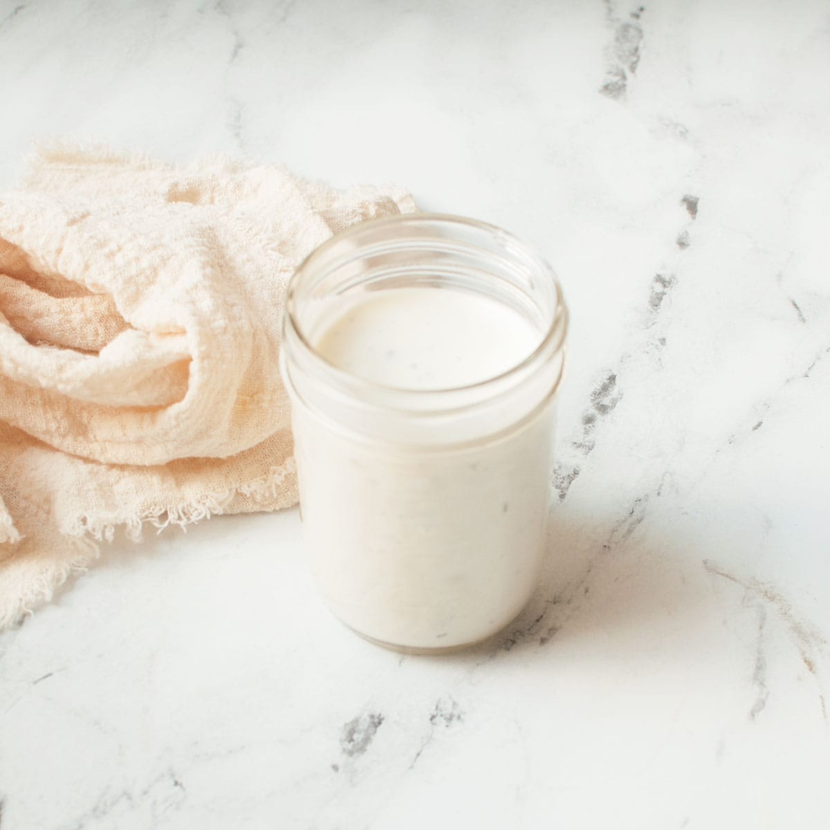 A photograph of copycat Creamy Parmesan Salad Dressing in a mason jar with a cream linen on a marble background.