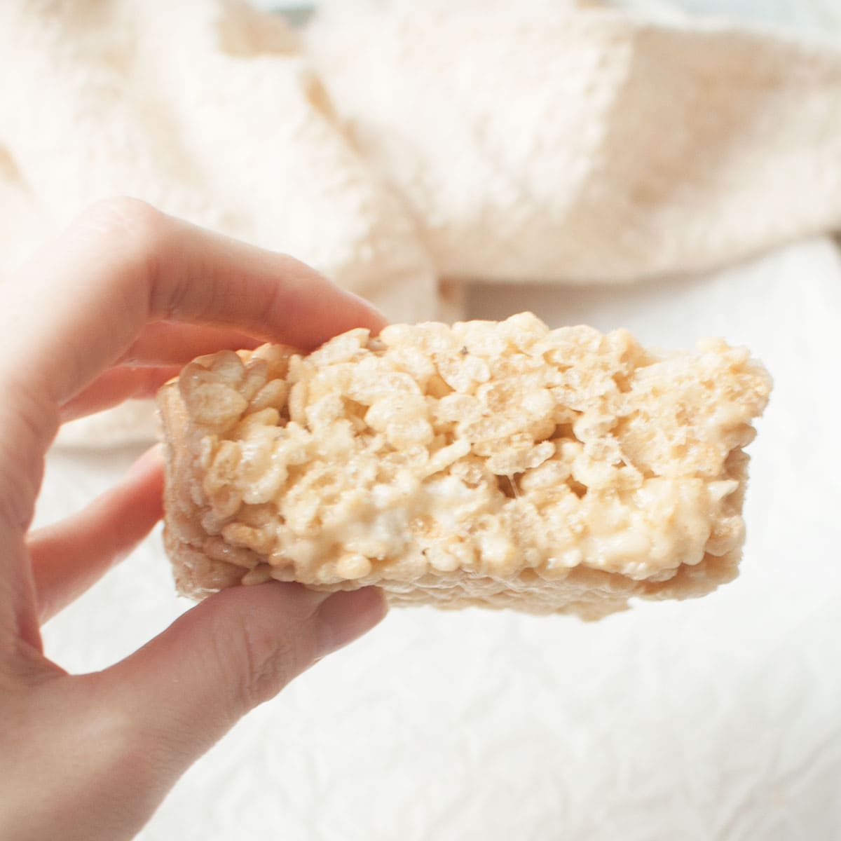 A hand holding a rice krispie treat. There's a marble background and a cream colored linen.